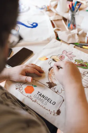 Hands decorating a Tangelo Games tote bag during a team workshop